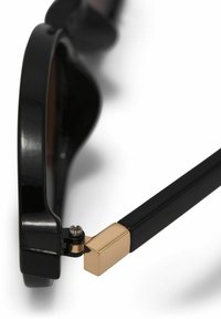 Monture de lunettes noire avec détail de charnière dorée reliant la branche à la monture avant, présentée sur un fond blanc.