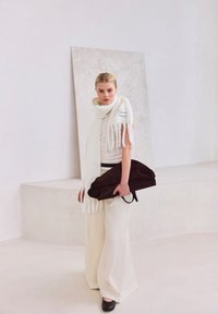 Witte gebreide sjaal met franjes, crème top en brede crème broek. Houdt een donkerbruine suède tas vast tegen een lichte neutrale achtergrond.