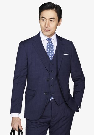 Mężczyzna w granatowym trzyczęściowym garniturze, białej koszuli i niebieskim krawacie w wzory, trzymający czarną torbę, patrzący na bok na białym tle.