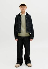 Garçon portant une casquette beige, un pull vert clair à plusieurs couches, une chemise blanche, une veste et un pantalon en denim foncé, ainsi que des chaussures beige, debout devant un fond blanc.