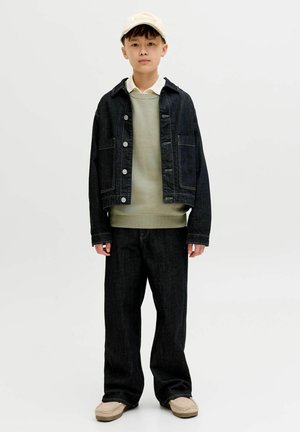 Poiss, kes kannab beežikat mütsi, rohelise toonides kihilist džemprit, valget särki, tumeda teksajahte ja -pükse ning beežeid kingi, seisab valge taustaga ees.