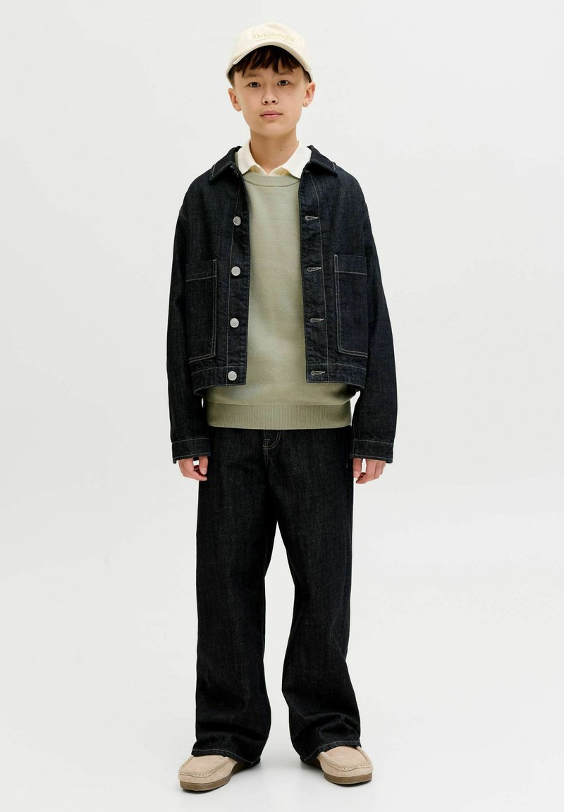 Garçon portant une casquette beige, un pull vert clair à plusieurs couches, une chemise blanche, une veste et un pantalon en denim foncé, ainsi que des chaussures beige, debout devant un fond blanc.