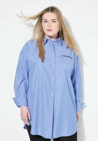 Camicia a righe blue e bianco con bottoni, maniche lunghe, taschino con la scritta "STUDIO UNITED" e vestibilità comoda. Tessuto morbido.