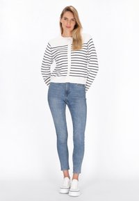Gestreifter weißer und navy-blauer Cardigan mit einer vorderen Knopfleiste, getragen über enganliegenden blauen Jeans und weißen Turnschuhen auf einem schlichten Hintergrund.