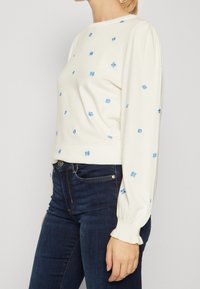 Vrouw in een witte lange mouwen trui met kleine blauwe bloemenborduursels, gecombineerd met donkerblauwe spijkerbroek, getoond van het midden van de romp tot de nek.