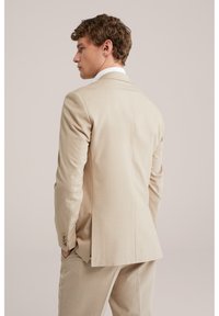 Uomo di profilo, indossa un giacca da completo color beige chiaro e pantaloni abbinati, con le mani in tasca su uno sfondo semplice.