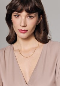 Złoty naszyjnik z łańcuszka o skręconym wzorze, noszony na szyi, na modelce w miękkiej beżowej bluzce z dekoltem w serek, na neutralnym tle.