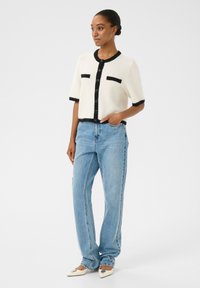White short-sleeve cardigan with black trim and pockets, paired with light blue straight-leg jeans and cream-colored pointed flats.