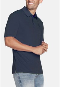 Mann, der lächelt, trägt ein dunkelblaues kurzärmeliges Polohemd und hellblaue Jeans, steht mit verschränkten Händen vor sich.