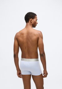 Bijele pamučne bokserice s zlatnim pojasom na kojem se nalazi logo "Calvin Klein", dizajnirane za usko pristajanje, s glatkom teksturom i oblikom.