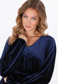 Tengerészkék bársonyblúz V-nyakkal, hosszú ujjakkal és begyűrt mandzsettákkal; puha tapintású és légies kialakítású, finom csillogással.