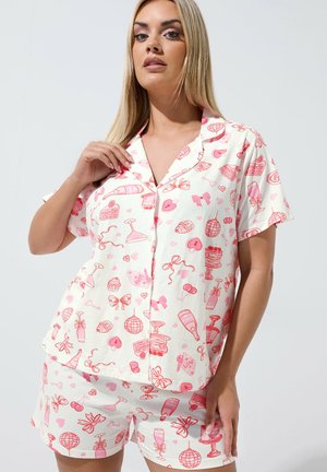 Nainen, joka pukeutunut valkoiseen, lyhythihainen nappipaitaan ja shortseihin, joissa on punaisia ja vaaleanpunaisia juhlateemaisia kuoseja, seisoo yksinkertaista taustaa vasten.