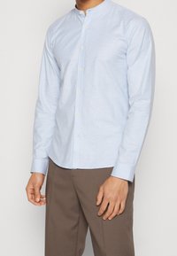 Camisa de manga larga de color azul claro con cuello de banda, botones frontales y una textura suave, combinada con pantalones marrones.