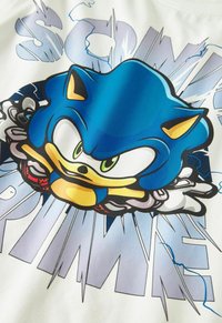 Hvid t-shirt med et blåt tegneserie-rygradsgrafik med grønne øjne, gule accenter og en eksplosiv baggrundstekst i nuancer af blåt.