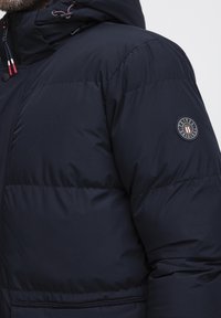 Marineblå polstret jakke med hætte, der har et rundt logo på ærmet, snoretræk og en glat ydre tekstur.