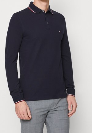 Mand iført navyblå poloshirt med lange ærmer og røde og hvide detaljer på krave og manchetter, kombineret med grå ternede bukser.