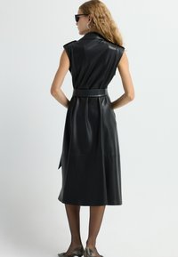 Zwarte faux leren jurk met een mouwloos ontwerp, kraaghals, belted taille en een vloeiende midi-lengte. Gladde textuur en minimale details.