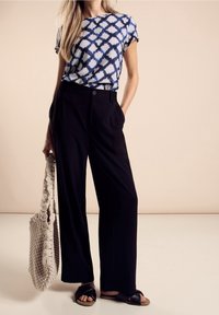 Blå och vit mönstrad kortärmad skjorta, högmidjade svarta vida byxor och svarta slidesandaler; håller en beige virkad toteväska.