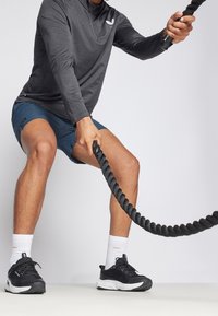 Musta pitkähiha paita, siniset shortsit ja mustat lenkkitossut valkoisin yksityiskohdin. Henkilö käyttää paksua mustaa taisteluköyttä aktiivisessa asennossa.