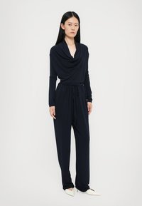 Navy blue jumpsuit with long sleeves, cowl neck, and drawstring waist. Fabric appears soft and drapes loosely. Model wears white pointed flats.
