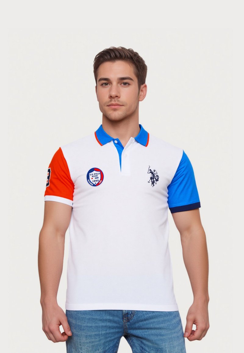 Joven con polo blanco con mangas rojas y azules, cuello azul, logotipos en el pecho y la manga, combinado con vaqueros azules.