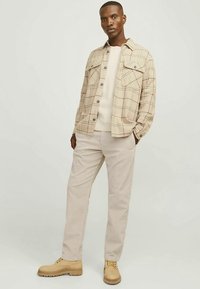 Jachetă cămașă bej cu carouri și buzunare la piept, pulover crem, pantaloni din catifea bej deschis și bocanci căpșunii cu șireturi. Design simplu și structurat.