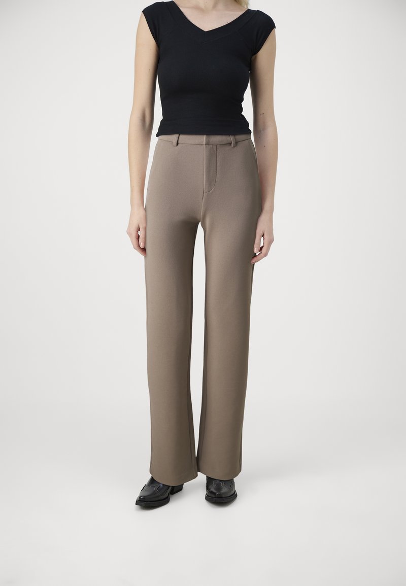 Femme portant un haut noir à manches courtes, un pantalon large taille haute coloris taupe, et des chaussures noires pointues, debout devant un fond uni.