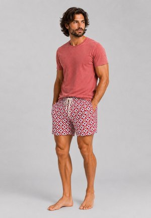 Mann steht barfuß, trägt ein ausgebleichtes rotes T-Shirt und rot-weiß gemusterte Shorts, mit den Händen in den Taschen, schaut zur Seite.
