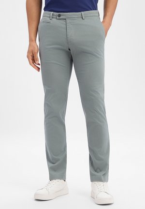 Mann trägt hellgraue, eng anliegende Chinos, navyblaues Hemd und weiße Sneaker, steht mit einer Hand in der Tasche.