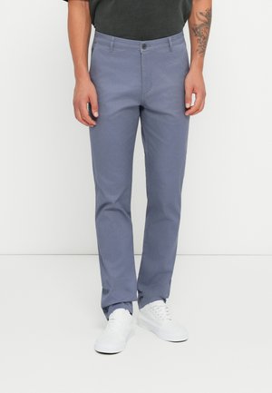 Chinos gris claro hechos de una mezcla de algodón con un corte slim. Cuenta con un cierre de botón y bolsillos frontales. Llevados con zapatillas blancas.