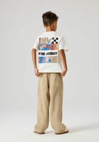 Garçon vu de dos portant un tee-shirt blanc avec des blocs graphiques multicolores et le texte « The Journey », un pantalon large beige, tenant l'ourlet de sa chemise.