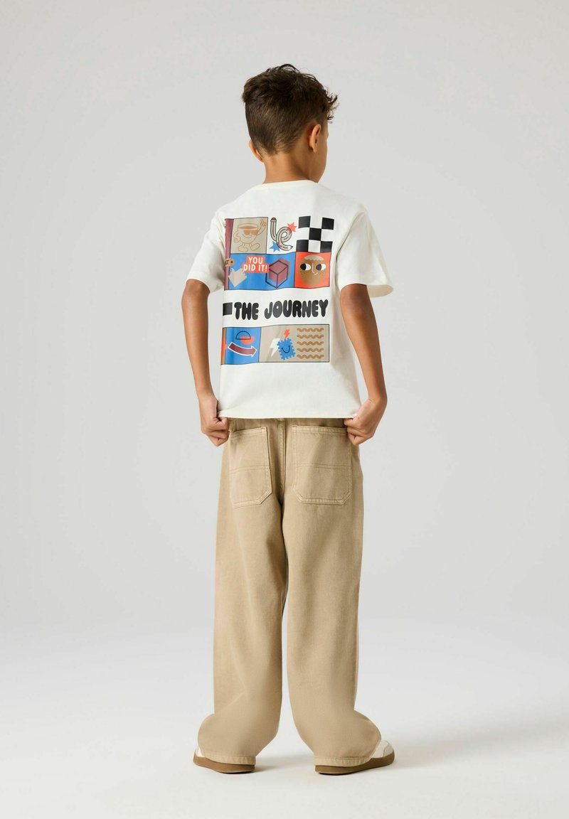 Garçon vu de dos portant un tee-shirt blanc avec des blocs graphiques multicolores et le texte « The Journey », un pantalon large beige, tenant l'ourlet de sa chemise.