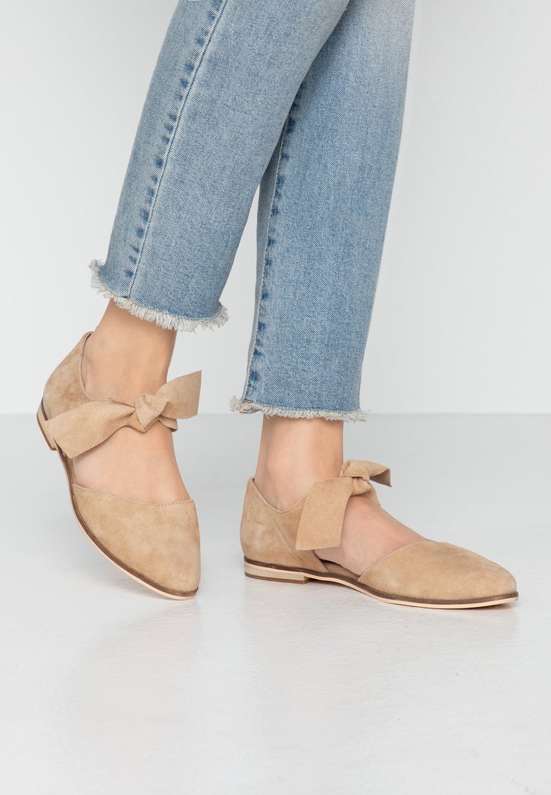 Pieds portant des chaussures plates en daim beige avec des nœuds, associées à un jean bleu clair à ourlet effiloché sur un sol blanc lisse.