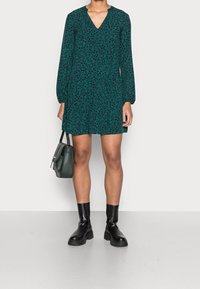 Grön V-ringad klänning med lång ärm och blommönster. Bärs med svarta ankelstövlar och en liten grön axelväska.