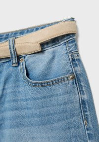Jean en denim bleu clair avec poche avant et ceinture en daim beige passée dans les passants, sur fond blanc.