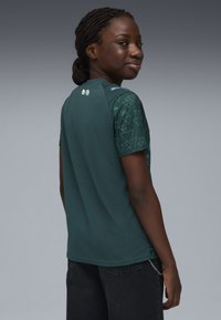 Teal színű sportpóló, rövid ujjakkal, geometrikus zöld mintákkal a vállakon és sima felülettel. A hátoldalon két gomb látható a gallérnál.