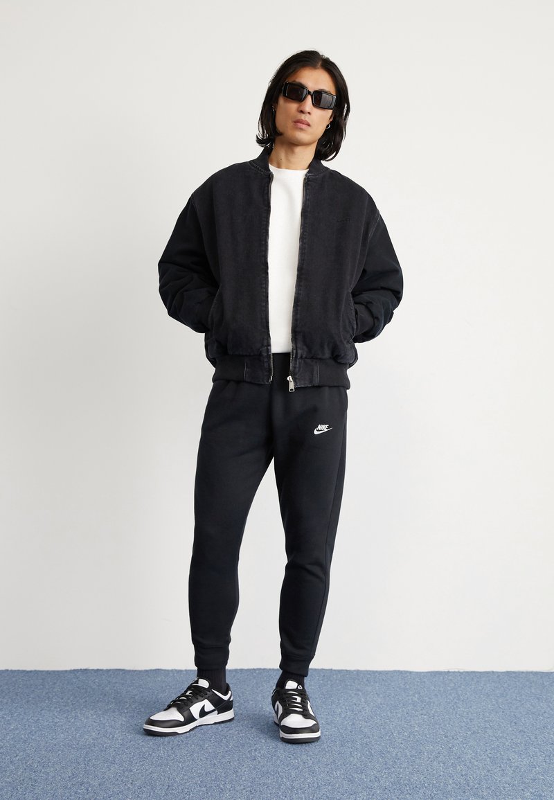 Chaqueta bomber negra con una tela ligeramente texturizada, camiseta blanca de cuello redondo, pantalones de chándal negros con el logo de Nike y zapatillas negras y blancas.