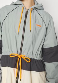 Chaqueta ligera con capucha en gris, con cordones naranjas y cintura ajustada. Bloques de color negro y crema en contraste completan el diseño.