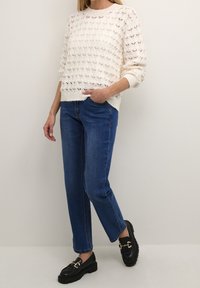 Jersey blanco de punto con un patrón texturizado, combinado con jeans rectos azules y mocasines negros con acentos de hardware dorado.