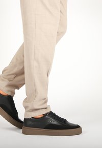 Sneakers in pelle nera e suede con suole in gomma marrone, caratterizzate da lacci neri e accenti minimi, abbinate a pantaloni beige chiaro.