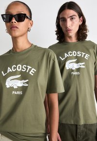 Olivno zeleni bombažni majici z belim logotipom Lacoste in grafiko krokodila, kratkimi rokavi, okroglo ovratnikom in sproščenim krojem.