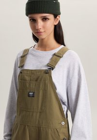 Olive green cotton overalls with adjustable straps, front pocket, and Vans logo; paired with a light gray long-sleeve shirt and dark green beanie.