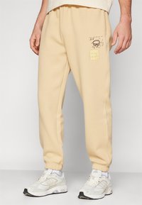 Beige sweatpants van een zachte stof, met een elastische tailleband en mouwen, voorzien van een grafisch ontwerp met de tekst "RENE DID IT FIRST" aan de zijkant.