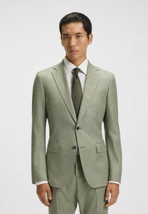 Man wearing light green suit with white shirt and dark green polka dot tie, standing against plain light background.