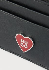 Portefeuille en cuir texturé noir arborant un logo en forme de cœur rouge et blanc avec le texte « HUGO ». Design simple avec des détails de couture visibles.