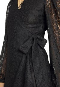 Gros plan sur une robe portefeuille en dentelle noire avec des manches longues transparentes et un grand nœud latéral, portée avec des colliers dorés superposés.