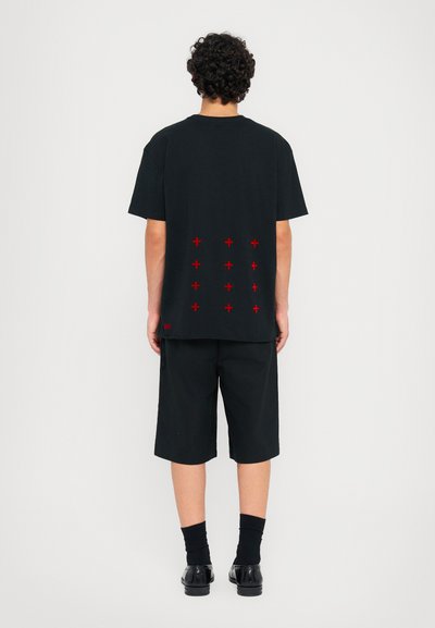T-shirt en coton noir à manches courtes, avec des motifs de croix rouges au dos. Assorti à un short noir et à des chaussures noires.