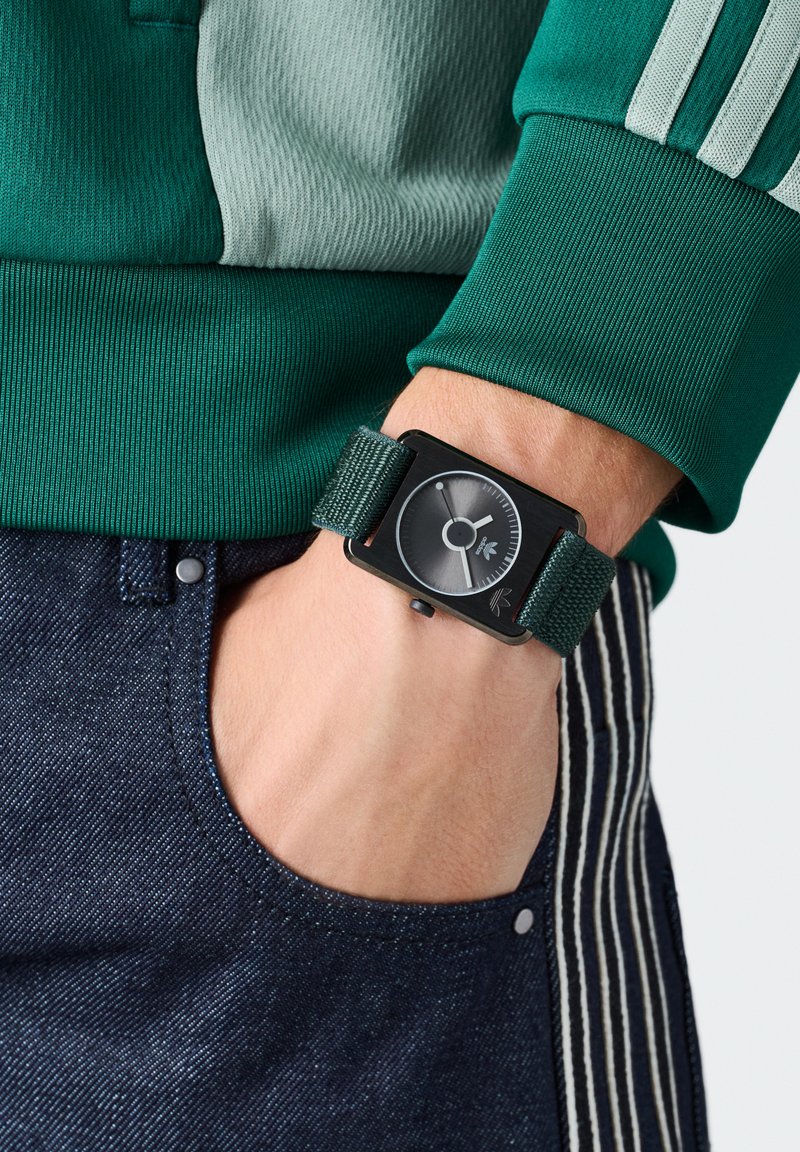 Montre rectangulaire noire avec un cadran gris, bracelet en tissu vert, portée sur une main. Jeans bleu foncé avec une poche et un bouton visibles dans le cadre.