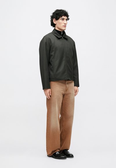 Čierna bunda na zips s vysokým golierom, v kombinácii s voľnými svetlohnedými nohavicami a čiernymi topánkami. Jednoduchý dizajn, textúra látky nie je detailne zaznamenaná.