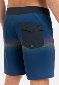 Shorts de bain bleus avec un motif à rayures horizontales dans des teintes de vert et de violet. Dotés d'une taille unie bleu foncé et d'une poche arrière.